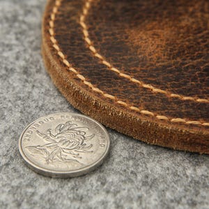 Puede incluir: Un posavasos de cuero marr&oacute;n con un borde cosido. El posavasos es redondo y tiene un aspecto desgastado. Una moneda china est&aacute; encima del posavasos.