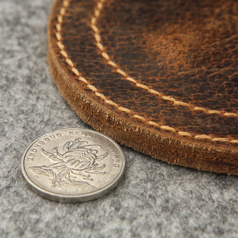 Puede incluir: Un posavasos de cuero marr&oacute;n con un borde cosido. El posavasos es redondo y tiene un aspecto desgastado. Una moneda china est&aacute; encima del posavasos.