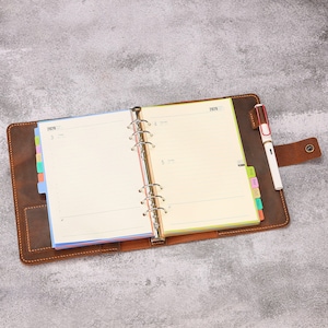 Op de afbeelding: Open bruin lederen planner met gelinieerde pagina's, gekleurde tabbladen en een pen. De planner is open op een spread met de datum 2026 zichtbaar. De pen is wit met een rode dop. Op een grijze ondergrond.