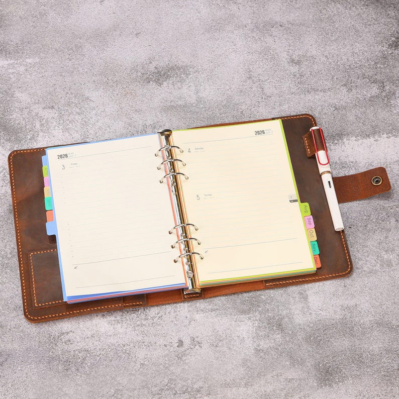 May include: A brown leather binder with a six-ring metal mechanism holding cream-colored 2026 planner pages. The planner features colorful side tabs, a snap closure, and a white and red fountain pen secured in a leather loop on the right side.