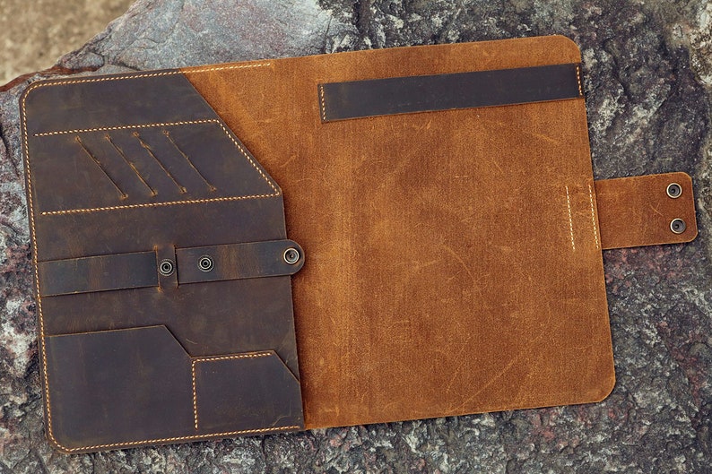 May include: A brown leather folder with multiple pockets and a flap closure. The folder is open and the inside is a lighter brown leather.
