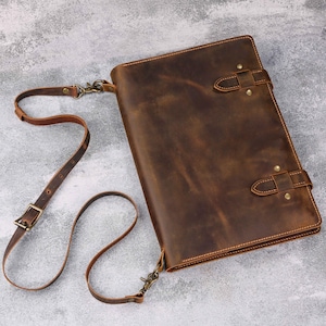 May include: A brown leather portfolio with a shoulder strap. The portfolio has two buckle closures and brass-colored rivets. The leather has a distressed appearance, and the strap is adjustable with a metal buckle.