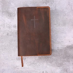May include: A brown leather Bible with a cross embossed on the cover. The word "James" is embossed at the bottom. The edges are stitched with a contrasting thread, and a brown ribbon bookmark is visible.