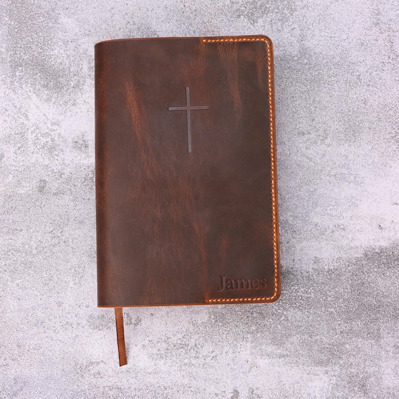 May include: A brown leather Bible with a cross embossed on the cover. The word "James" is embossed at the bottom. The edges are stitched with a contrasting thread, and a brown ribbon bookmark is visible.