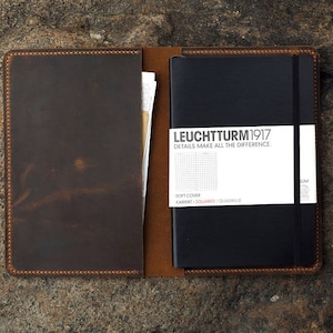 Puede incluir: Una cubierta de cuaderno de cuero marr&oacute;n con un cuaderno negro en el interior. El cuaderno tiene el texto "LEUCHTTURM1917" y "DETAILS MAKE ALL THE DIFFERENCE" en la portada.