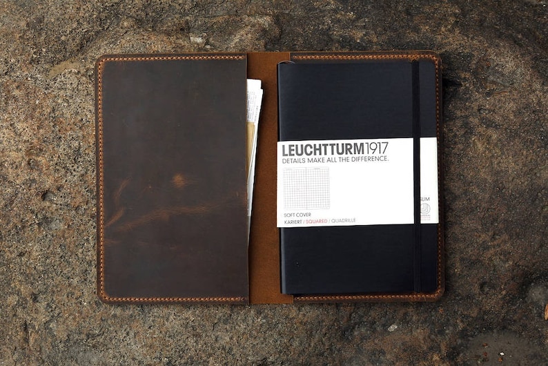 Puede incluir: Una cubierta de cuaderno de cuero marr&oacute;n con un cuaderno negro en el interior. El cuaderno tiene el texto "LEUCHTTURM1917" y "DETAILS MAKE ALL THE DIFFERENCE" en la portada.
