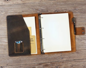 Könnte beinhalten: Offener brauner Lederordner mit einem Stift und einer Tasche für Karten. Der Ordner hat eine leere weiße Seite und eine Seite mit dem Text "Field Notes" darauf.