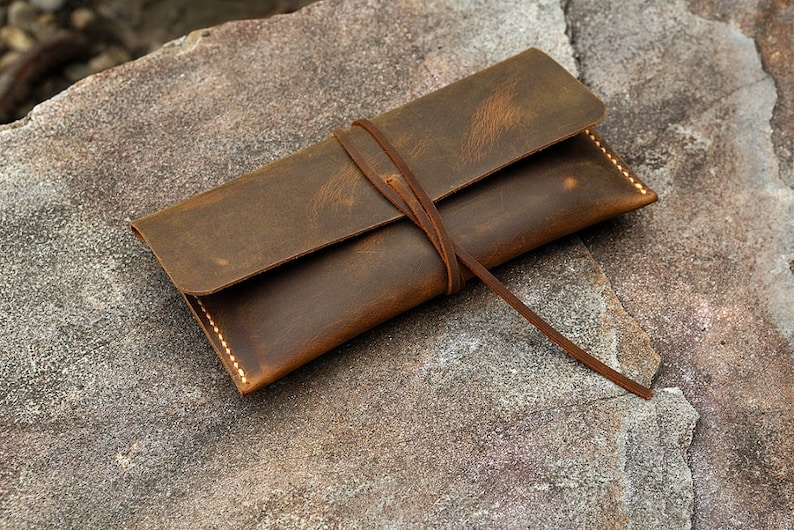 Op de afbeelding: Een bruine leren potlood etui met een strik sluiting. De etui heeft een verweerde look en is gemaakt van dik, duurzaam leer.