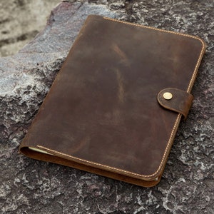 May include: A brown leather notebook with a brass button closure. The notebook is lying on a rough, gray rock surface.
