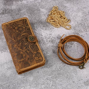 May include: A brown leather wallet with a floral embossed design and a brass button closure. The wallet is accompanied by a brown leather strap and a gold chain.
