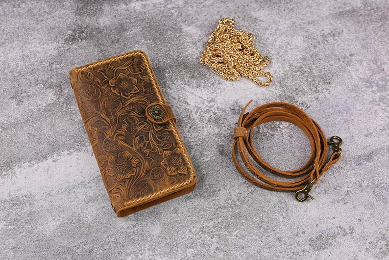 May include: A brown leather wallet with a floral embossed design and a brass button closure. The wallet is accompanied by a brown leather strap and a gold chain.