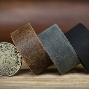 以下が含まれることがあります： 茶色、灰色、黒の革ひも3本。ひもは巻かれて木製の表面に置かれています。前景には「YES」と書かれた小さなコインがあります。