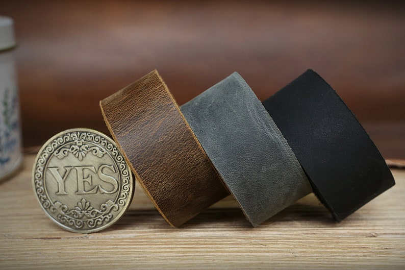 以下が含まれることがあります： 茶色、灰色、黒の革ひも3本。ひもは巻かれて木製の表面に置かれています。前景には「YES」と書かれた小さなコインがあります。