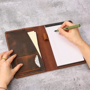 Puede incluir: Una funda de cuero marrón oscuro para cuaderno con un bolsillo vertical, una ranura pequeña para tarjetas y una trabilla para bolígrafo. La funda contiene un bloc de notas blanco con líneas y una pluma estilográfica verde salvia.