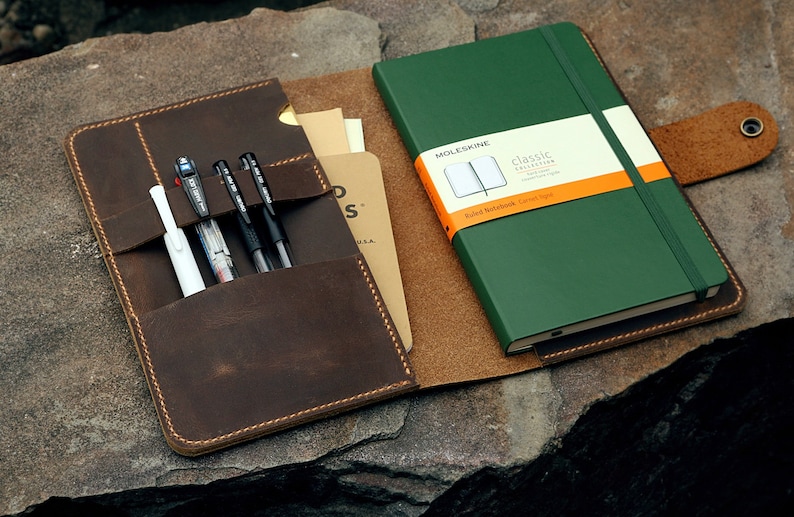 Op de afbeelding: Een bruine leren notitieboekhoes met een groen Moleskine notitieboek en verschillende pennen. De hoes heeft vakken voor pennen en een knoopsluiting. Het notitieboek heeft een wit en oranje label.