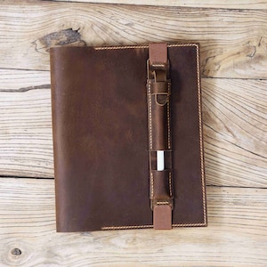 May include: A brown leather notebook cover with a pen holder and a stitched edge. The cover is on a light wood surface.
