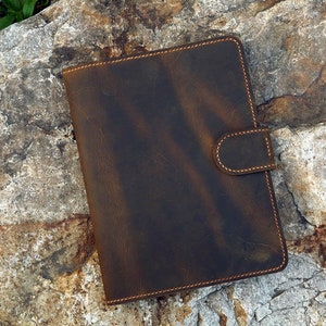 May include: A brown leather notebook with a flap closure and a stitched edge. The notebook is sitting on a rough, gray rock.