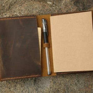 Puede incluir: Un cuaderno de cuero marrón con un portaplumas y una página en blanco en el interior. El cuaderno tiene un borde cosido y una solapa que se cierra con un botón.