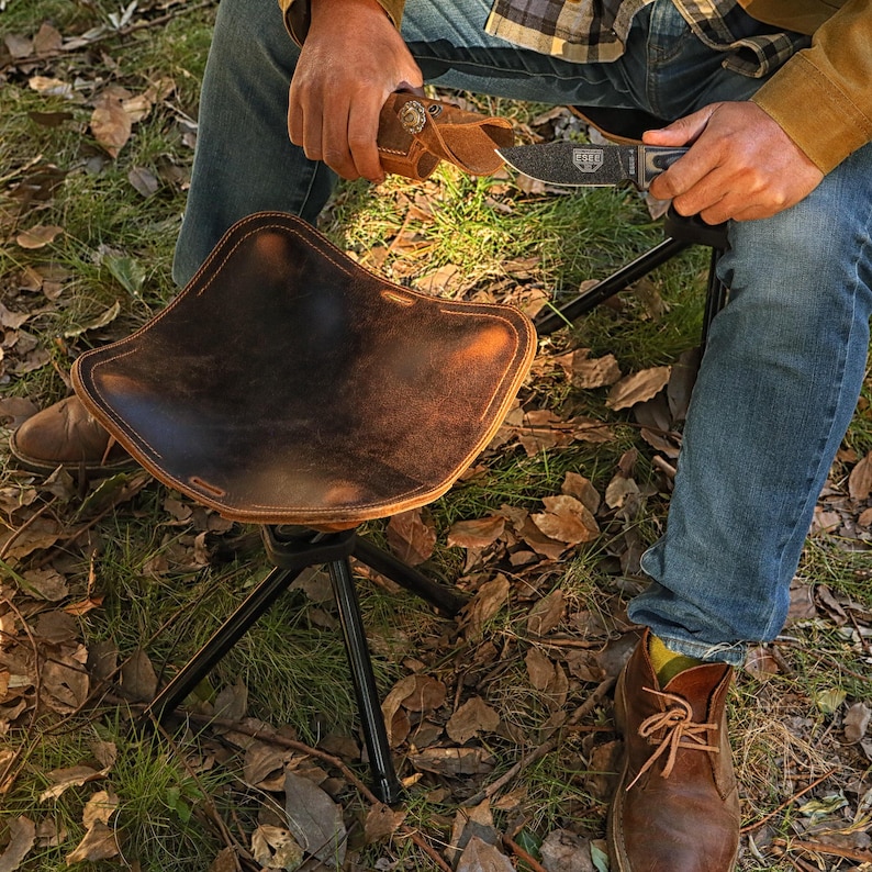Puede incluir: Una silla de camping de cuero marr&oacute;n con un marco de metal negro. La silla est&aacute; instalada en una superficie cubierta de hierba con hojas ca&iacute;das. Una persona est&aacute; sentada en la silla y sostiene un cuchillo.
