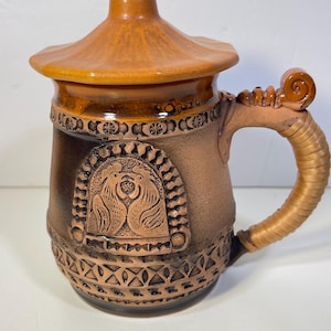 May include: A brown ceramic mug with a matching lid, featuring a relief design of two birds facing each other within an arched frame. The mug has a textured, wrapped-style handle and decorative geometric patterns around the base and rim.
