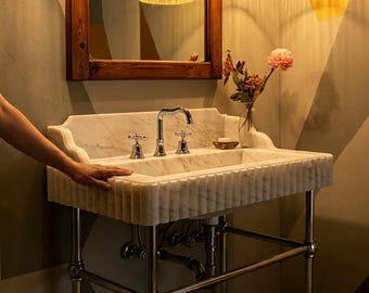 May include: A white marble bathroom sink with a fluted front edge and a high backsplash, supported by a polished chrome metal frame. The sink features a central chrome faucet with two cross-handle knobs and a clear glass vase with flowers.