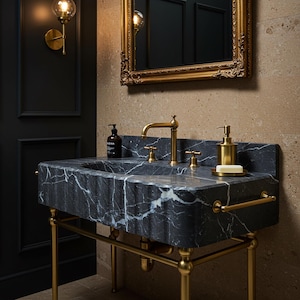 May include: A black marble bathroom sink with white veining, featuring a brass faucet, two brass handles, a brass towel bar, and a brass soap dispenser. The sink is supported by a brass metal frame and includes a small bar of soap on a tray.
