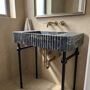 Op de afbeelding: Een donkergrijze marmeren wastafel met een geribbeld exterieurontwerp, ondersteund door een zwart metalen frame. De wastafel is voorzien van een aan de muur gemonteerde messing kraan en bijpassende messing leidingen onder het bekken, tegen een beige betegelde muur.