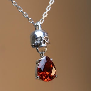 May include: A silver skull pendant necklace with a teardrop-shaped red gemstone hanging from it.