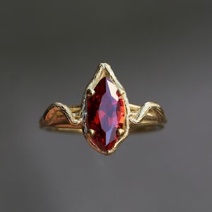 以下が含まれることがあります： 葉の形をしたデザインに赤い宝石がセットされた金の指輪です。