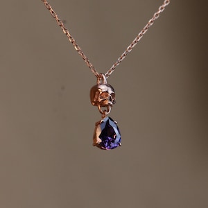 May include: A rose gold necklace with a skull pendant and a dangling purple gemstone.