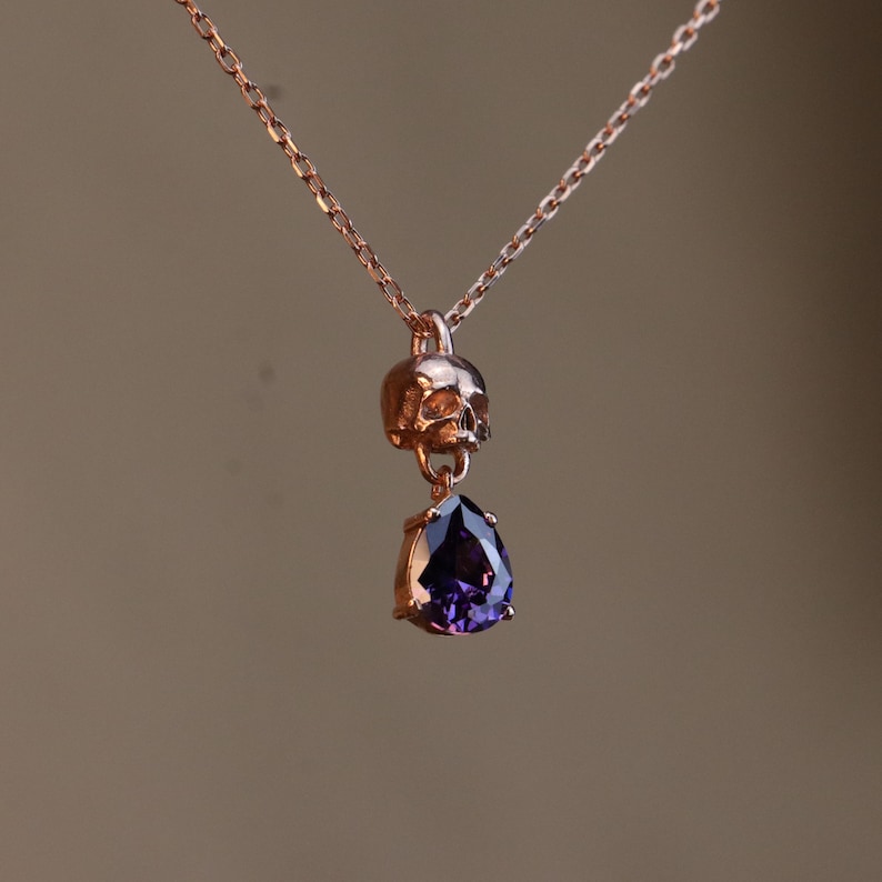 May include: A rose gold necklace with a skull pendant and a dangling purple gemstone.