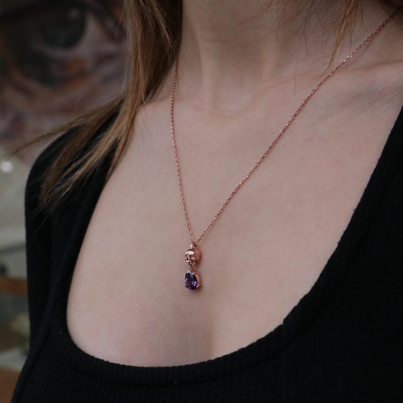 May include: A rose gold necklace with a small skull charm and a purple gemstone pendant.