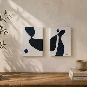 Puede incluir: Dos pinturas abstractas con formas azul marino sobre lienzo blanco. Una lámpara de cerámica, un cuenco pequeño y libros están sobre una superficie de madera. Un jarrón con vegetación está a la izquierda.