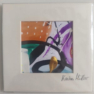 Puede incluir: Pintura abstracta en acuarela en un marco blanco. La obra presenta pinceladas naranjas, moradas, verdes y negras con salpicaduras blancas. Un pequeño objeto dorado está en la parte inferior. La firma del artista, "Karina Miller", está en la esquina inferior derecha.