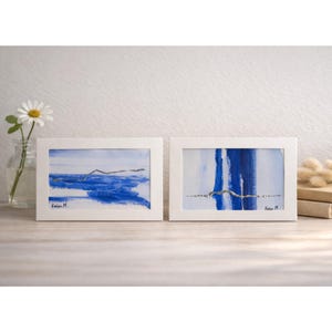 Puede incluir: Dos pinturas abstractas enmarcadas con marcos blancos. Las pinturas presentan pinceladas azules y blancas. Una pintura representa un paisaje con un mar azul y una fina línea dorada. La otra tiene trazos azules verticales.