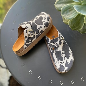 May include: A pair of black and white cow print slip-on clogs with a buckle detail. The clogs have a cork footbed and a rubber outsole.