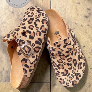 May include: Two leopard print clogs with cork soles. The clogs have a brown and tan pattern, with a buckle closure. The text "Anatomik Mantar Taban" is visible on a circular label. The clogs are on a wooden surface.