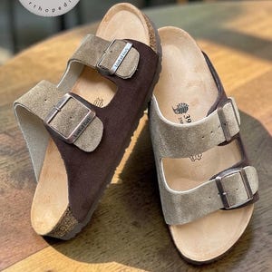 May include: Brown and beige sandals with buckle fastenings. The sandals feature a cork sole and a suede-like upper. The text "Anatomik Mantar Taban Orthopedic" is visible in the background.