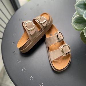 Puede incluir: Un par de sandalias de ante marrón con hebillas dobles y detalles dorados. Las sandalias tienen una suela de plataforma de corcho.
