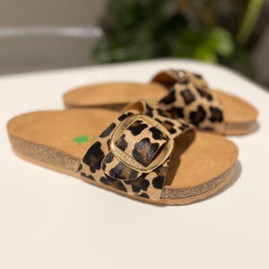 Pu&ograve; includere: Un paio di sandali marroni e neri con stampa leopardata e una fibbia dorata. I sandali hanno una piattaforma in sughero e un cinturino che passa sopra il piede.