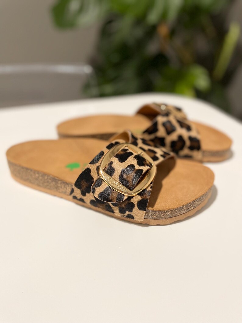 Pu&ograve; includere: Un paio di sandali marroni e neri con stampa leopardata e una fibbia dorata. I sandali hanno una piattaforma in sughero e un cinturino che passa sopra il piede.