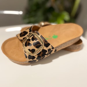 Pu&ograve; includere: Sandali con stampa leopardata, fibbia dorata e plateau in sughero. I sandali hanno un cinturino in pelle marrone con il nome del marchio "land of leather" stampato su di esso.