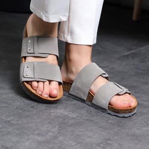 Op de afbeelding: Een paar grijze suède sandalen met twee verstelbare banden en een kurk voetbed.