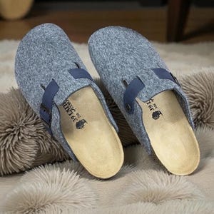 Puede incluir: Un par de zuecos de fieltro gris con correas y hebillas azul marino. Los zuecos tienen una plantilla clara y contorneada y descansan sobre una alfombra esponjosa de color crema. Los zuecos son de la talla 39.