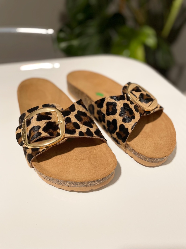 Pu&ograve; includere: Un paio di infradito con stampa leopardata e fibbia dorata. Le infradito hanno una piattaforma in sughero e un cinturino in pelle marrone.