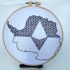 May include: A black and white embroidery of Antarctica with a geometric design in the center, displayed in a wooden embroidery hoop.
