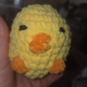 May include: A yellow crochet chick with an orange beak and feet. The chick has small black eyes and is made of soft yarn. This handmade plush toy is a cute and cuddly item.