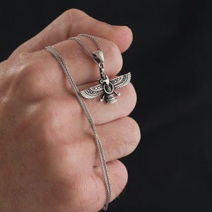 May include: A silver pendant necklace featuring a detailed Faravahar design. The pendant depicts a winged figure with a circular emblem, suspended from a delicate chain. The necklace is held against a dark background, highlighting the intricate details.