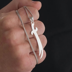 May include: A silver-coloured pendant shaped like a curved sword, known as a scimitar, suspended from a thin, woven silver-coloured chain. The pendant features a smooth, polished metallic finish with a distinct crossguard and a pointed, curved blade.