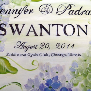 May include: A painted sign with the names "Jennifer & Padraic" and the last name "Swanton" in elegant script. The date "August 20, 2011" is also displayed, along with "Saddle and Cycle Club, Chicago, Illinois". The sign is decorated with watercolor-style blue and green hydrangeas.